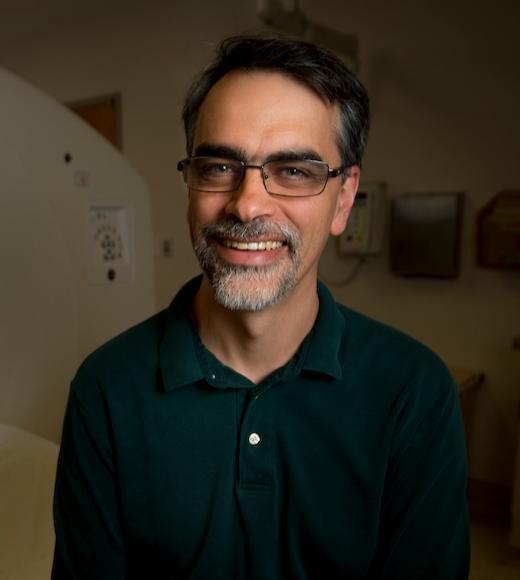 Dr. Badawi in dark polo shirt, glasses, smiling, scanner in background